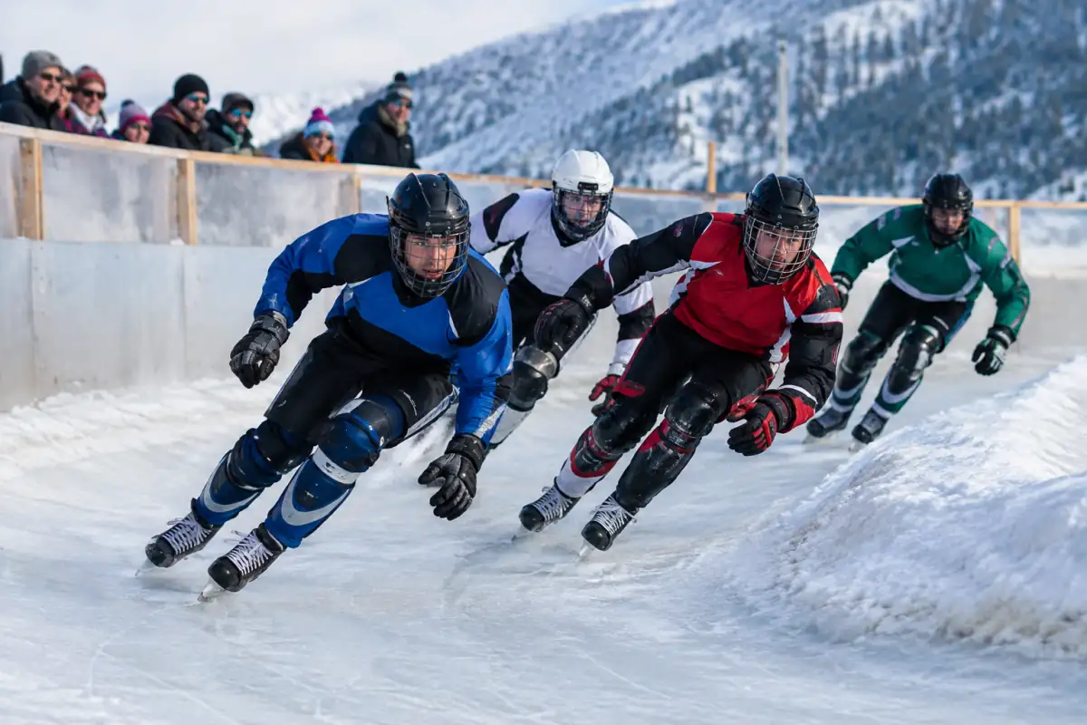 Quatre patineurs équipés de protections négocient un virage relevé sur une piste d’ice cross glacée en plein air.