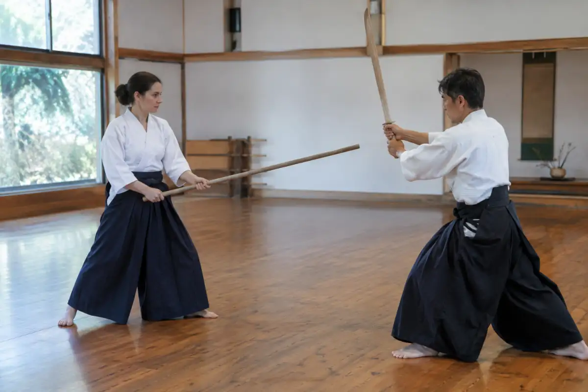 Deux pratiquants de jōdō répètent un kata au jō et au bokken dans un dojo, en contrôlant leurs gestes.