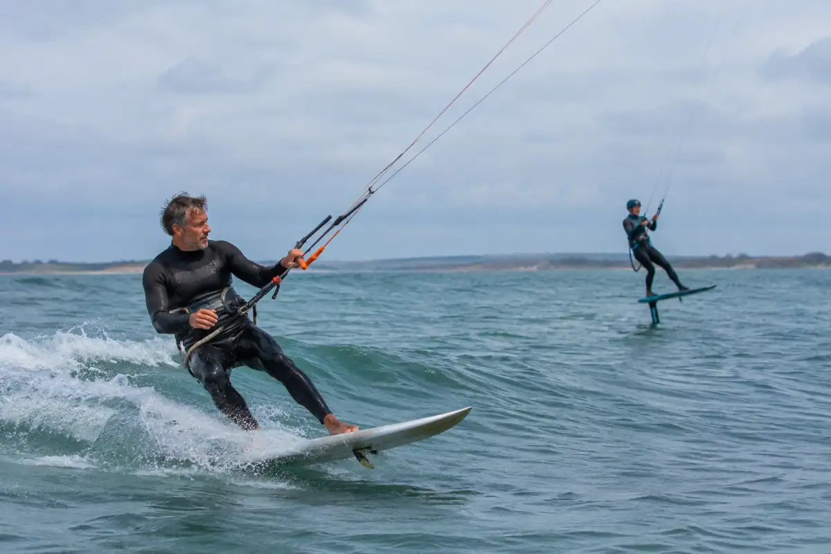 Un kitesurfeur sur planche directionnelle suit une petite vague tandis qu’un autre glisse au-dessus de l’eau sur un foil.