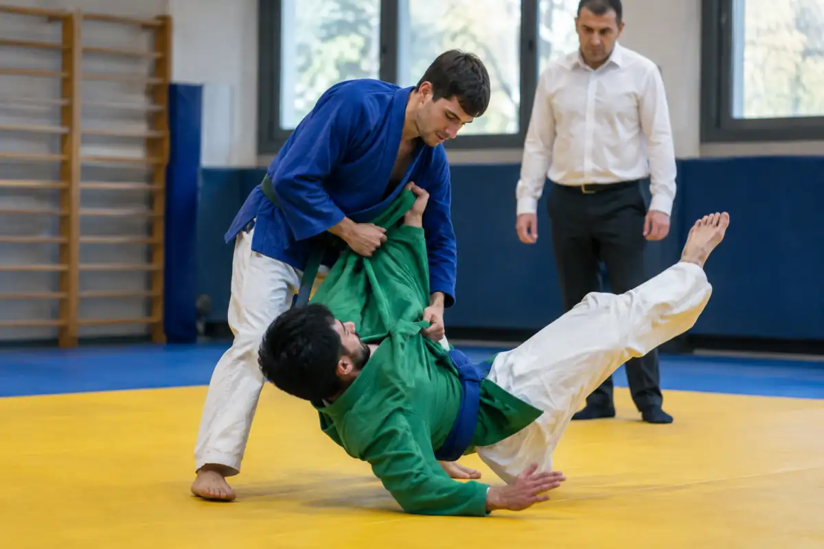 Deux adversaires en vestes bleue et verte exécutent une projection debout de kurash sur un tapis, sous la surveillance d’un arbitre.