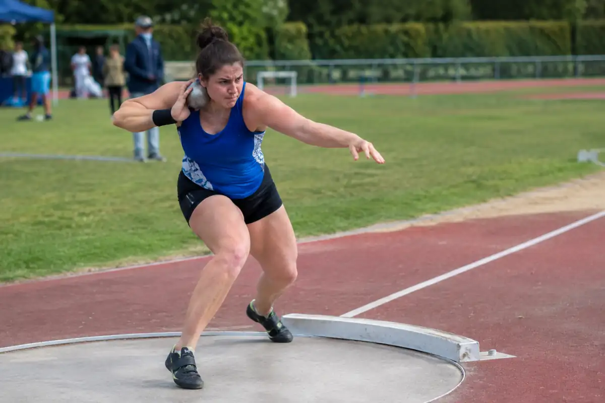 Athlète propulsant un poids métallique depuis le cercle lors d’un entraînement de lancer du poids sur un stade.