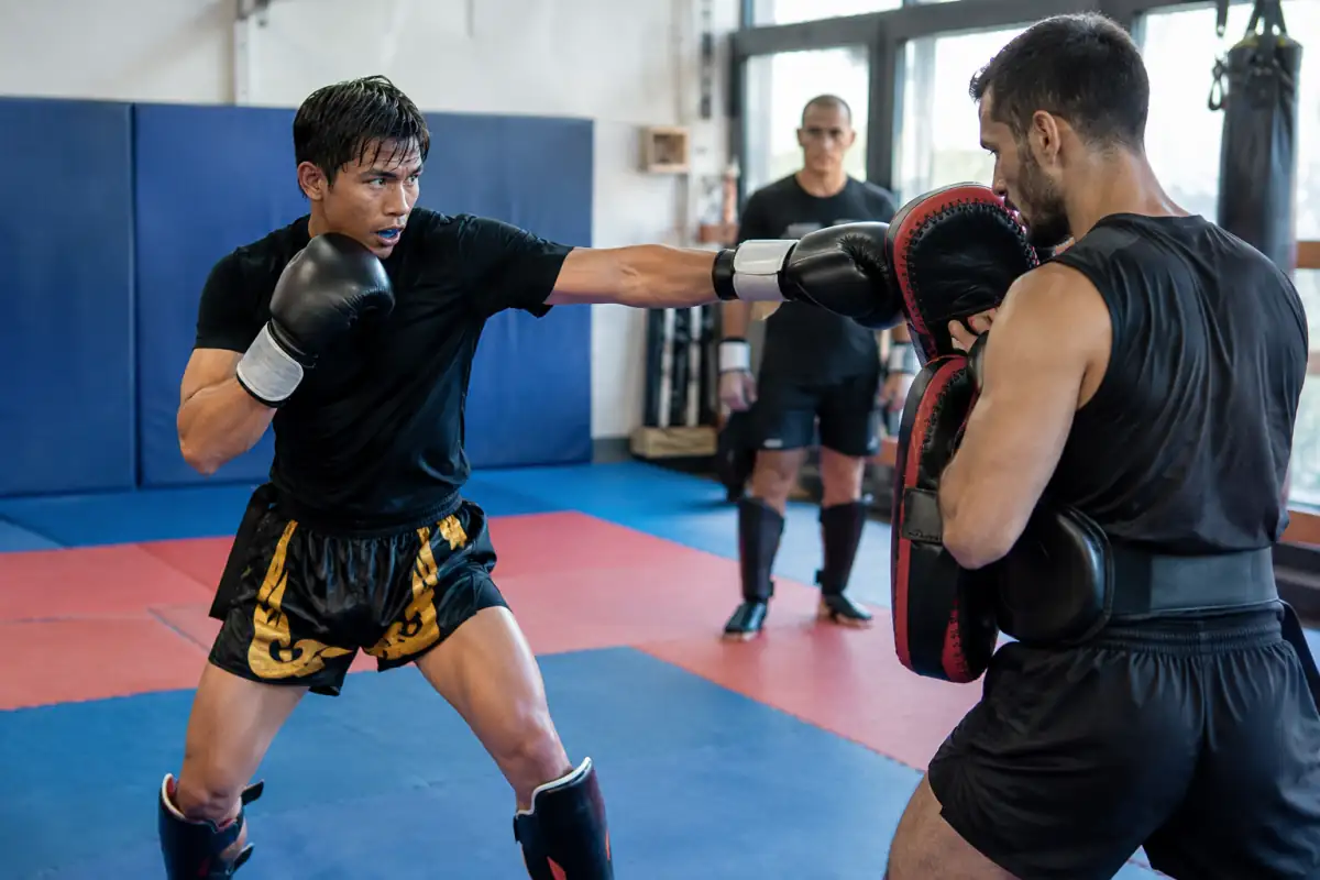 Deux pratiquants travaillent les frappes de lethwei sur paos, en gants et protège-tibias dans une salle de sport.