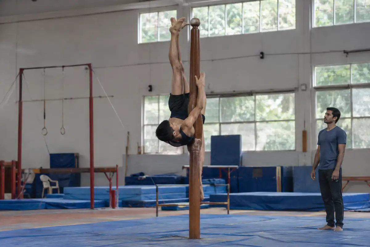 Athlète en position inversée sur un poteau de mallakhamb, au-dessus de tapis de réception dans une salle d’entraînement.