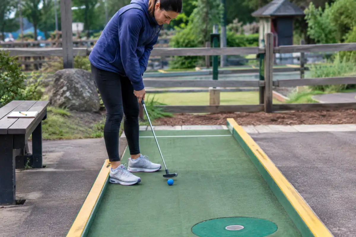 Joueur de mini-golf travaillant un putt droit sur une piste dégagée, avec une balle alignée vers la cible et un carnet de suivi à proximité.