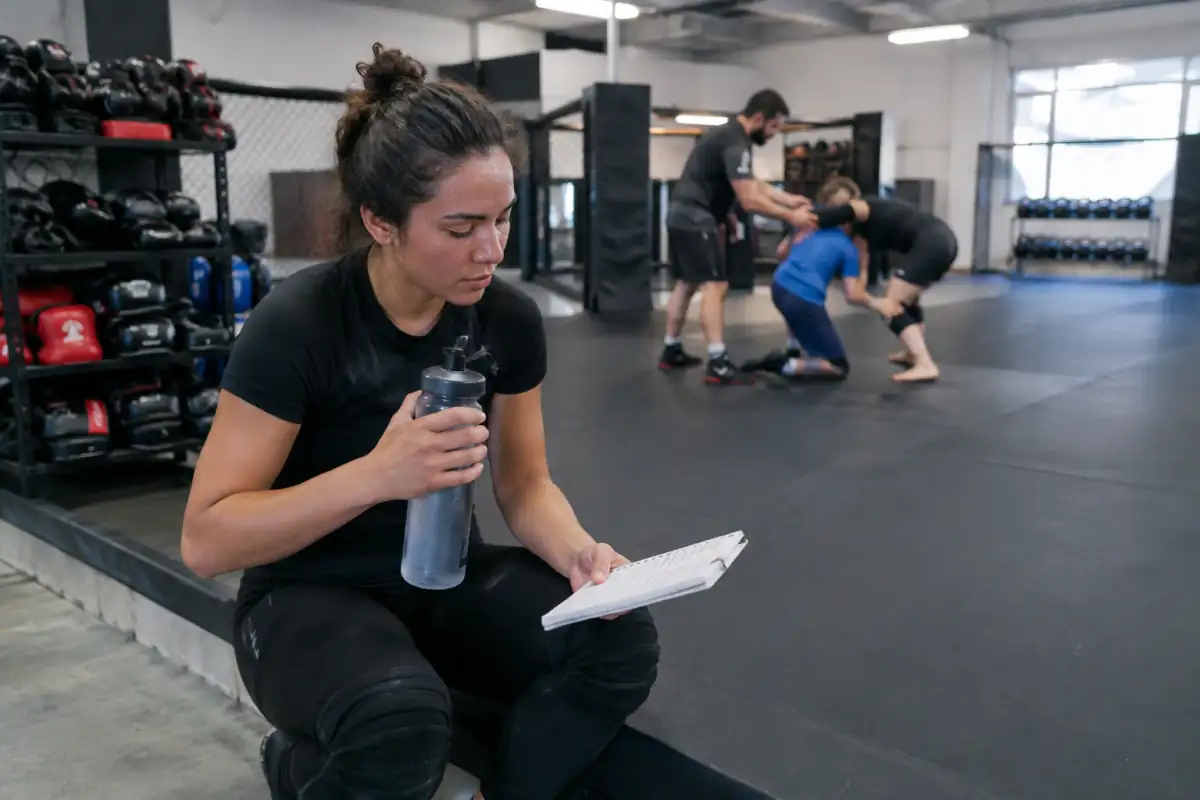 Un débutant en MMA récupère entre deux exercices pendant qu’un coach encadre un travail de lutte contrôlé sur les tapis.