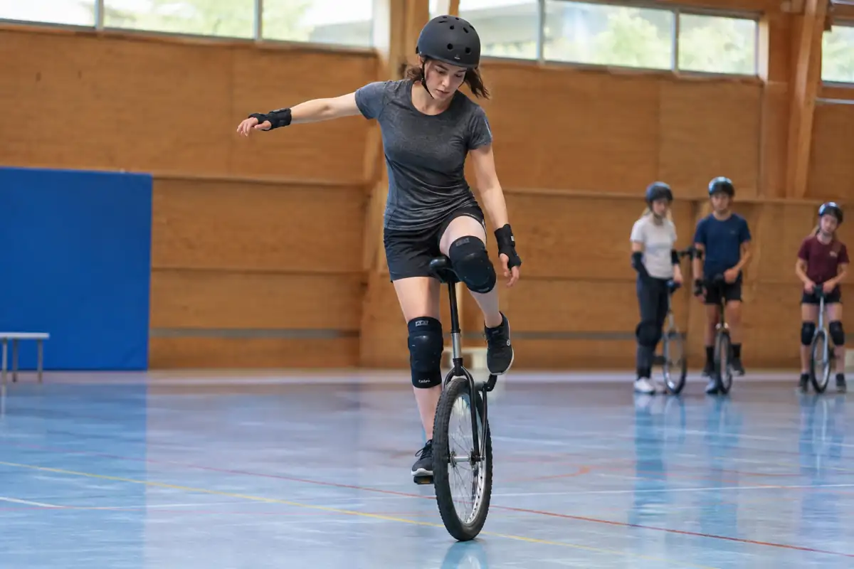 Une monocycliste enchaîne une figure de freestyle sur un monocycle artistique dans un gymnase, avec casque et protections.