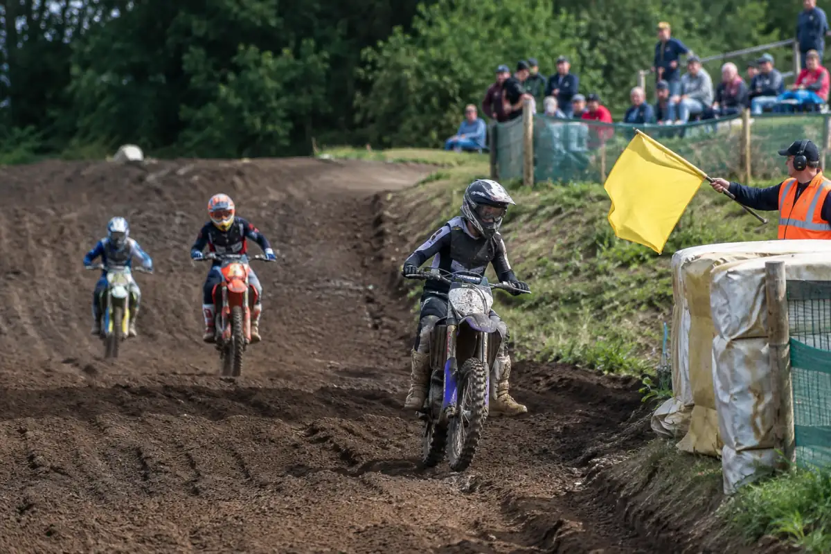 Un commissaire agite un drapeau jaune au bord d’une piste de motocross tandis que les pilotes ralentissent dans la zone de danger.