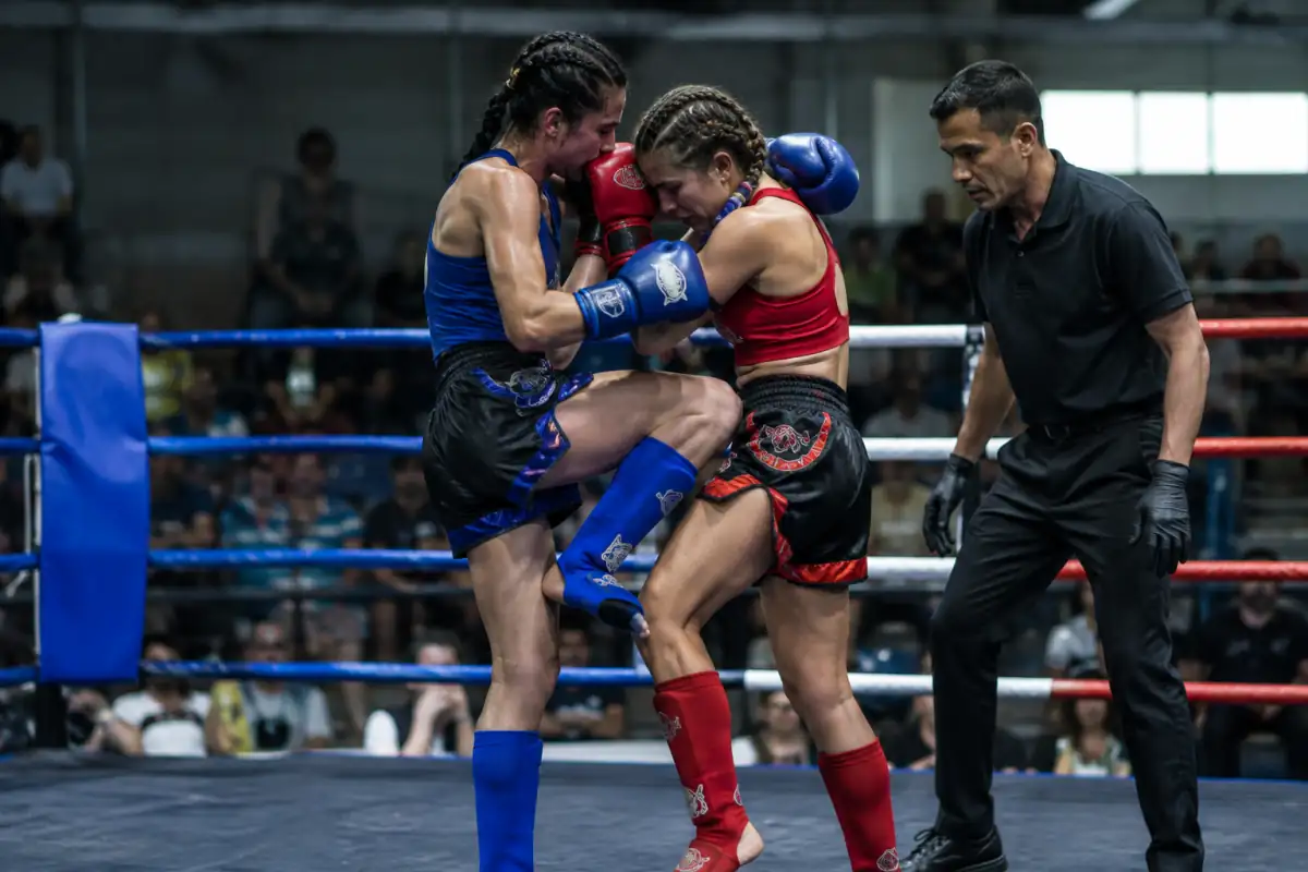 Deux combattants échangent un genou au corps en clinch dans un ring de muay-thaï, sous le contrôle attentif de l’arbitre.