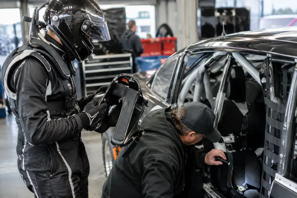 Pilote en combinaison vérifiant son casque et son système de retenue près d’une voiture de stock-car inspectée au garage.