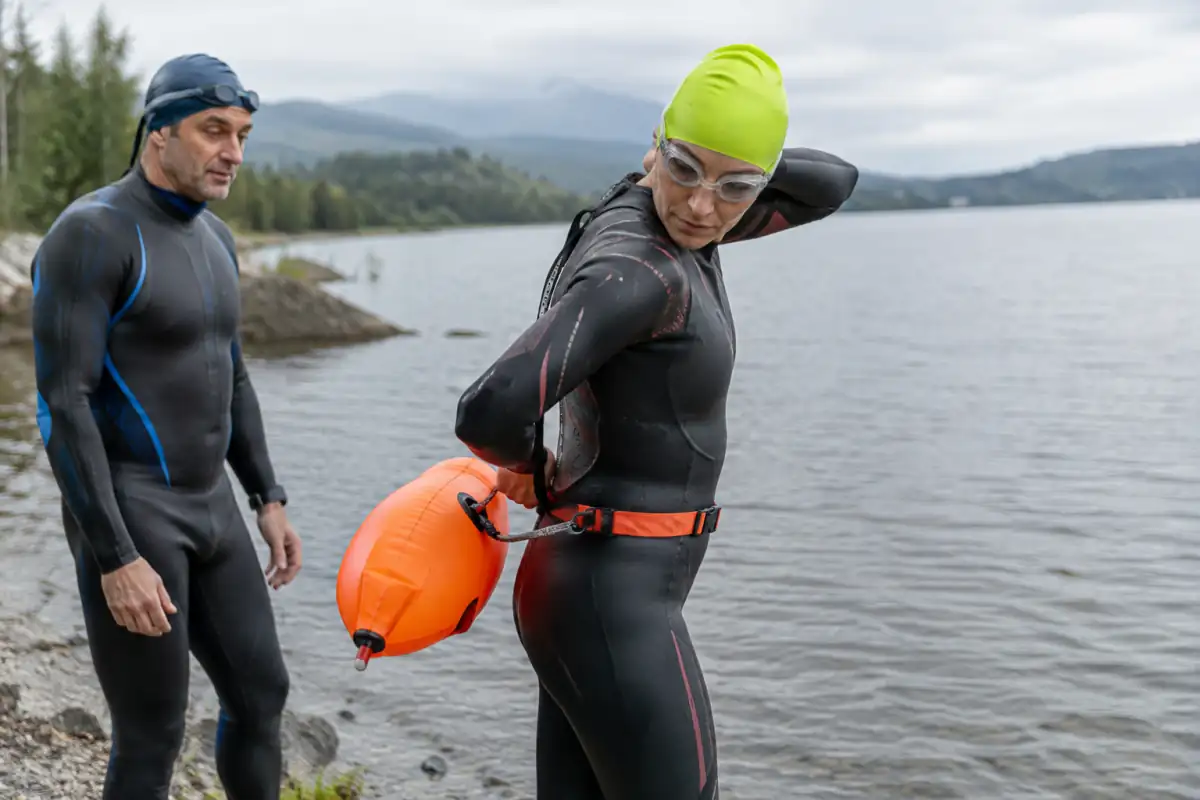 Nageur en combinaison ajustant la sangle d’une bouée orange au bord d’un lac, bonnet vif et lunettes adaptés à l’eau libre.