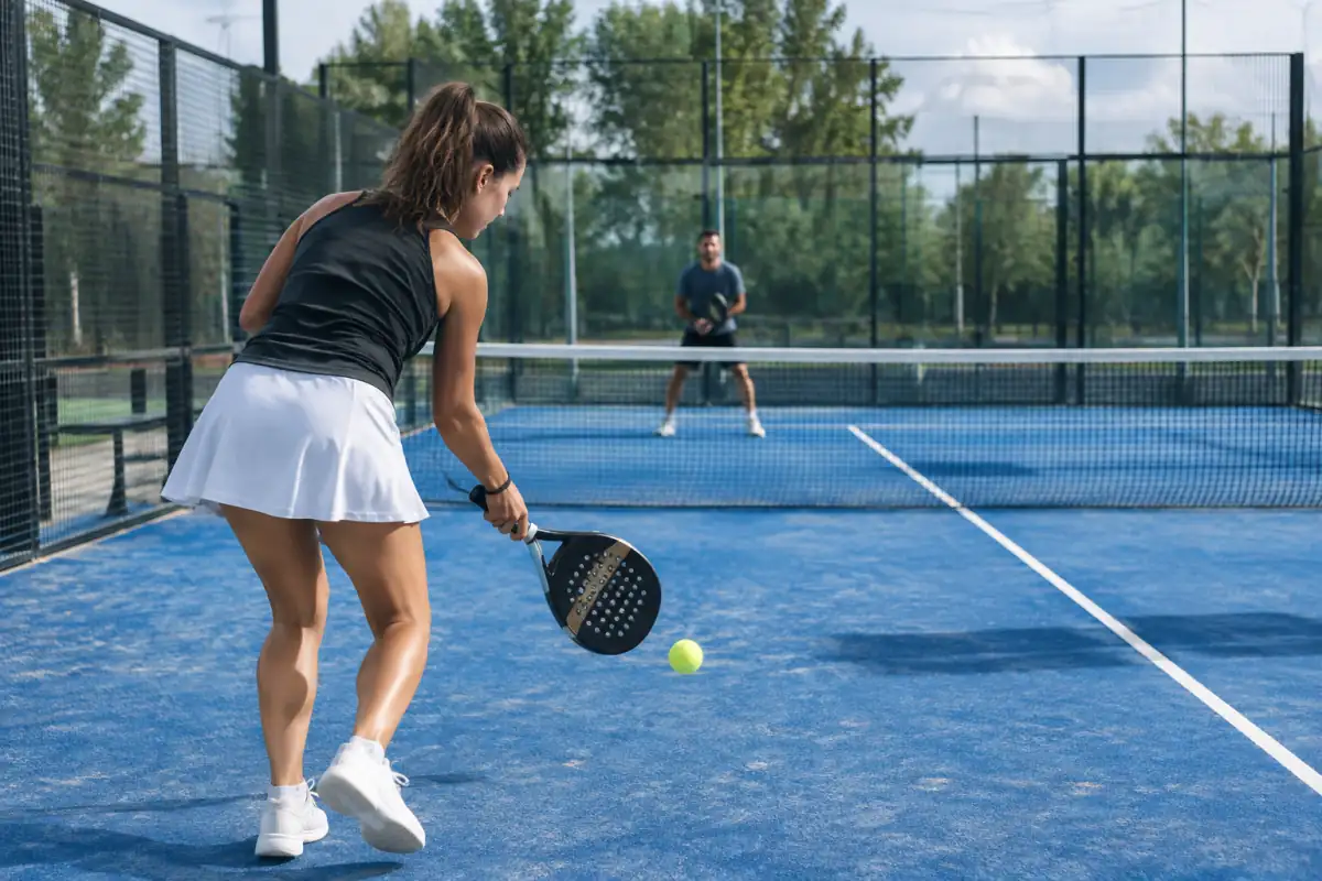 Joueur de padel préparant un service à la cuillère derrière la ligne, avec le filet, le carré diagonal et la vitre visibles.
