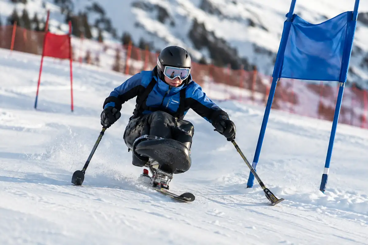 Un skieur en monoski adapté enchaîne un virage entre les portes d’un parcours de para ski alpin en montagne.