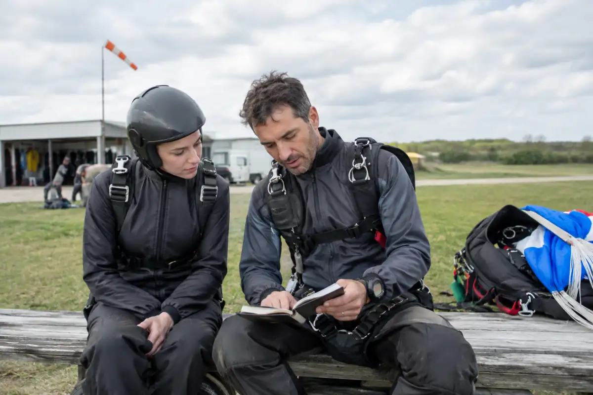 Un parachutiste débutant échange avec son instructeur après un saut et note un objectif de progression dans son carnet.