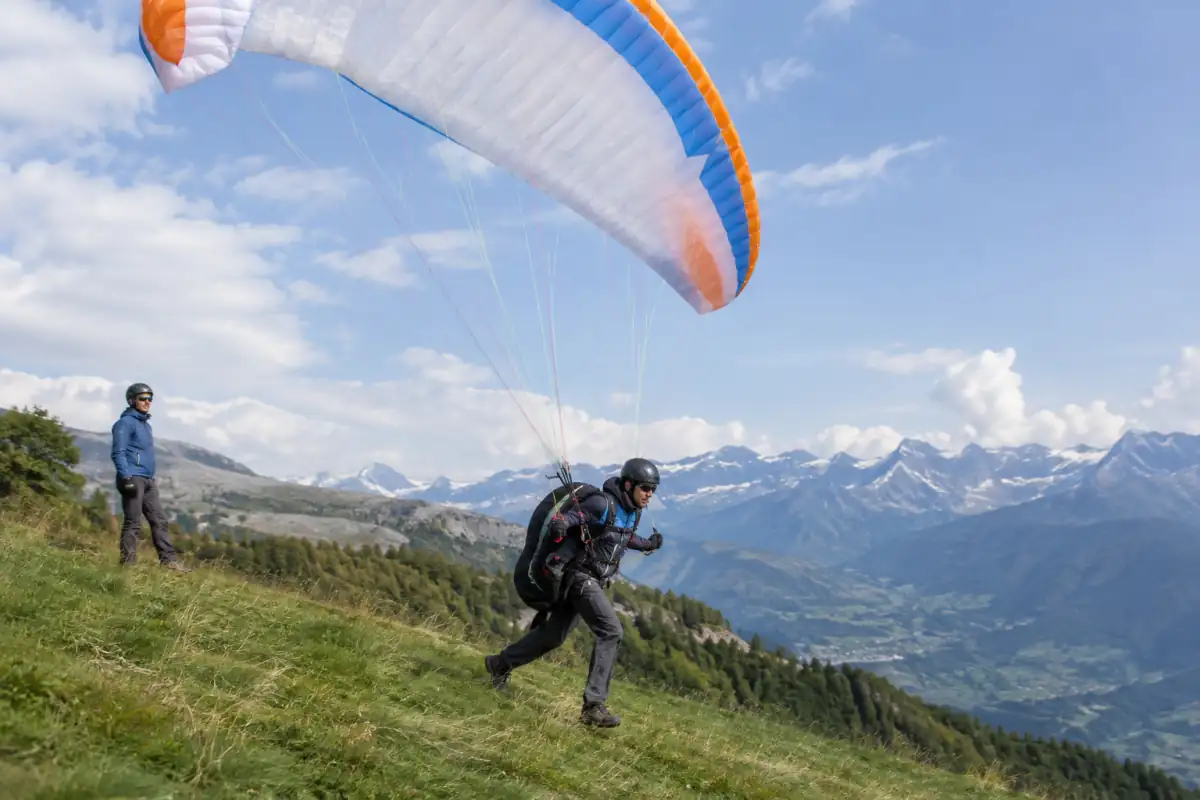 Un pilote décolle à pied en parapente sur une pente alpine, sous son aile souple et équipé d’une sellette et d’un casque.
