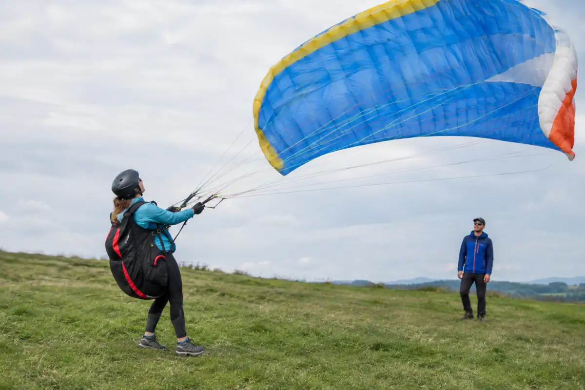 Un débutant travaille le gonflage de sa voile sur une pente école, sous le regard d’un moniteur de parapente.