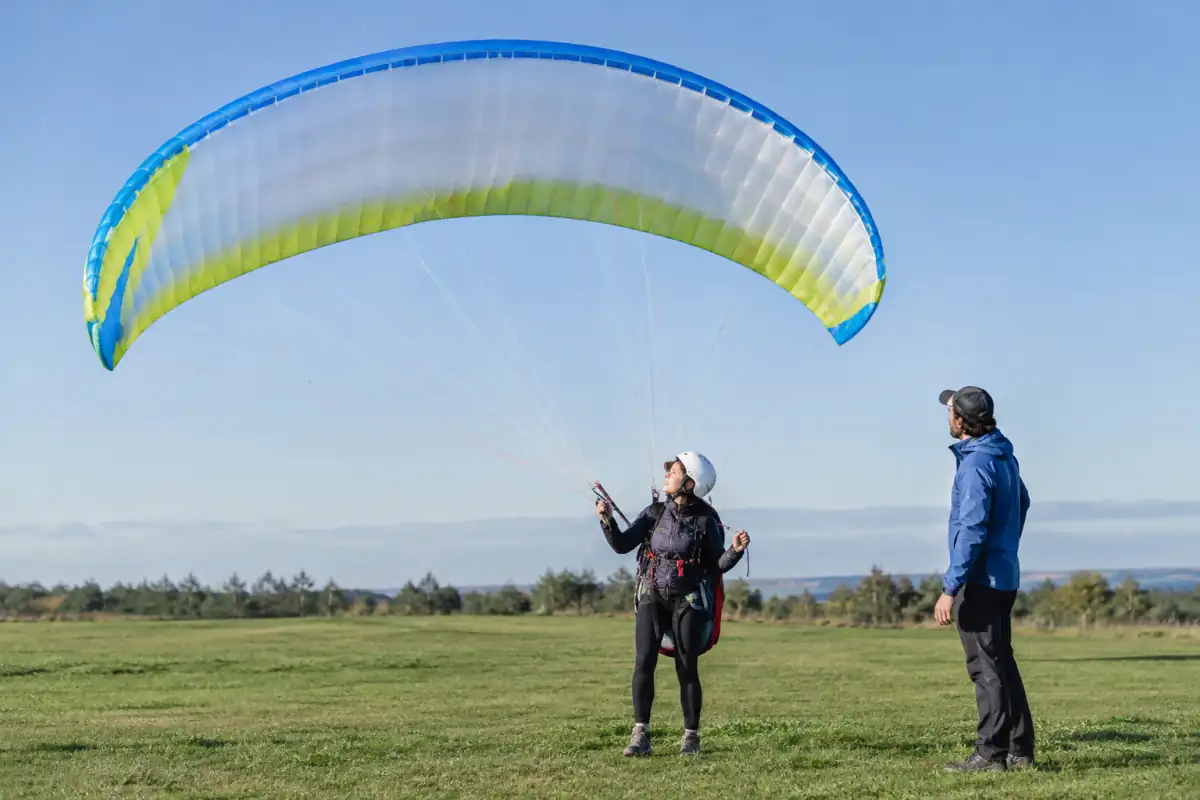Un débutant stabilise sa voile de parapente au sol pendant un exercice de gonflage supervisé sur un terrain dégagé.