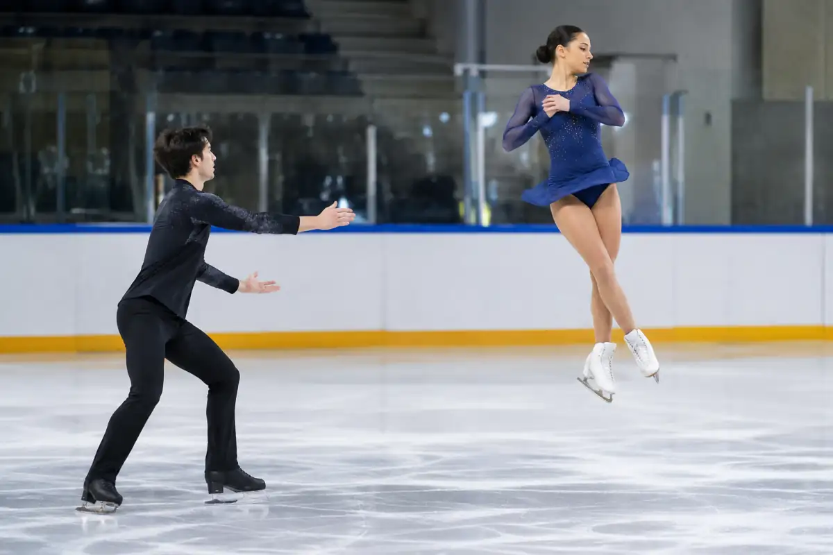 Deux patineurs réalisent un saut lancé en couple sur la glace d’une patinoire couverte, sous l’œil de leur entraîneur.