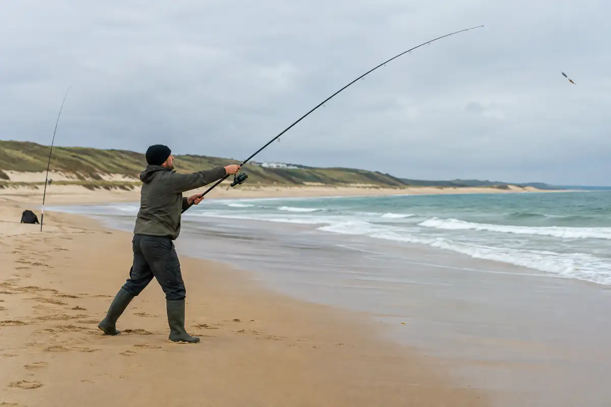 Un pêcheur lance sa canne de surfcasting depuis une plage de la côte atlantique, face aux vagues.