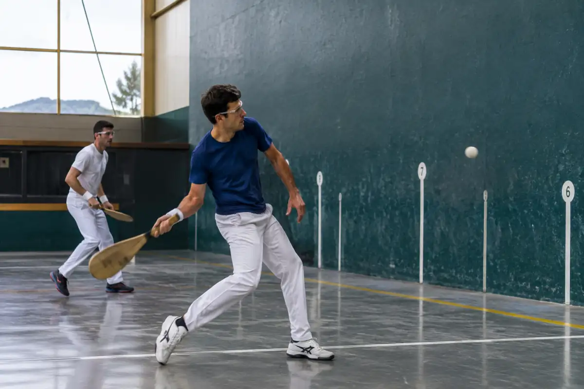 Joueurs de pelote basque en échange dans un fronton couvert, l’un frappe la pelote avec une pala tandis que l’autre se replace.