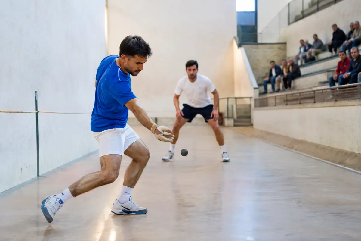 Joueurs de pelote valencienne échangeant une balle à main protégée dans un trinquet, au-dessus de la corde centrale.