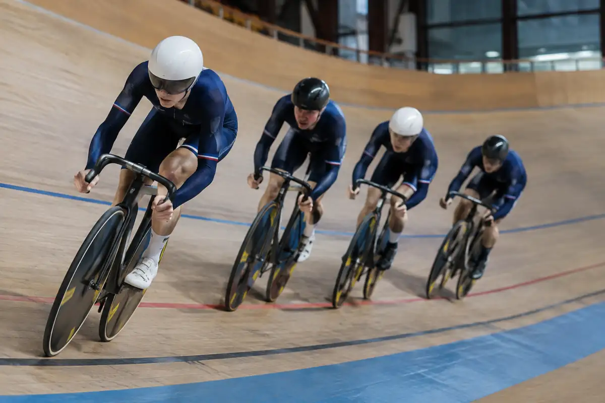 Quatre cyclistes en file réalisent une poursuite par équipes sur la piste relevée d’un vélodrome couvert.