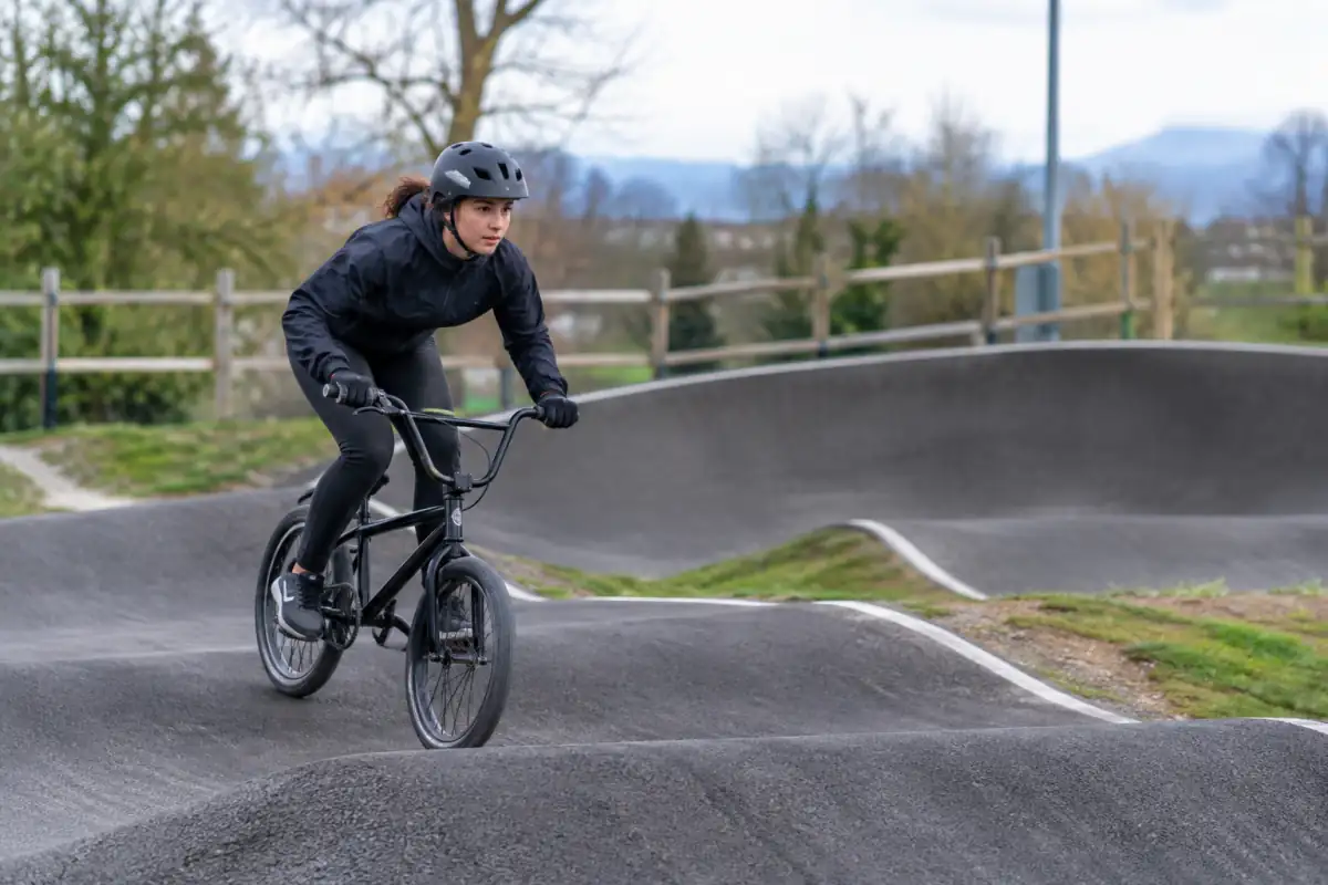 Un cycliste en BMX pompe sur une bosse d’un pumptrack en enrobé, casque ajusté et regard tourné vers le virage relevé.