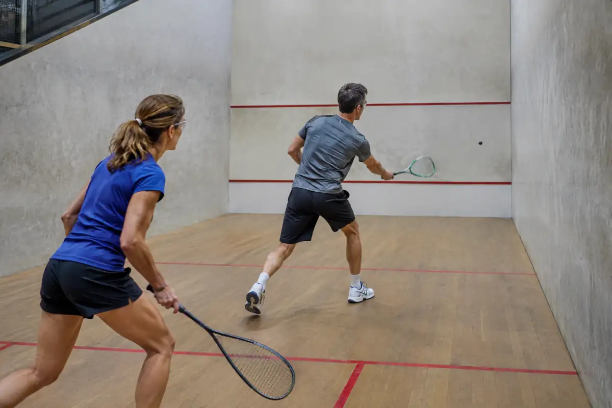 Deux joueurs disputent un échange de rackets dans un court fermé, raquettes levées face au mur frontal.