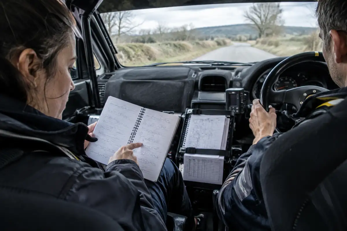 Un copilote de rallye suit ses notes manuscrites pendant une reconnaissance, tandis que le pilote reste attentif à la route.
