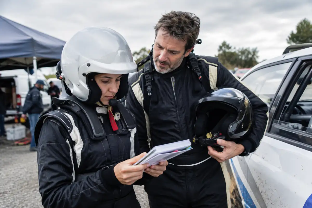 Un copilote débutant vérifie son carnet de route et son casque près d’une voiture de rallye à l’arrêt.