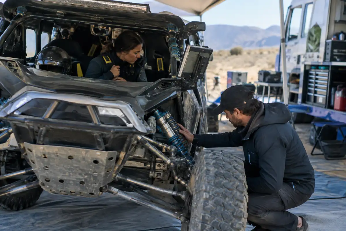 Équipage vérifiant le harnais, l’intercom et le matériel de navigation d’une voiture de rallye-raid avant une étape.