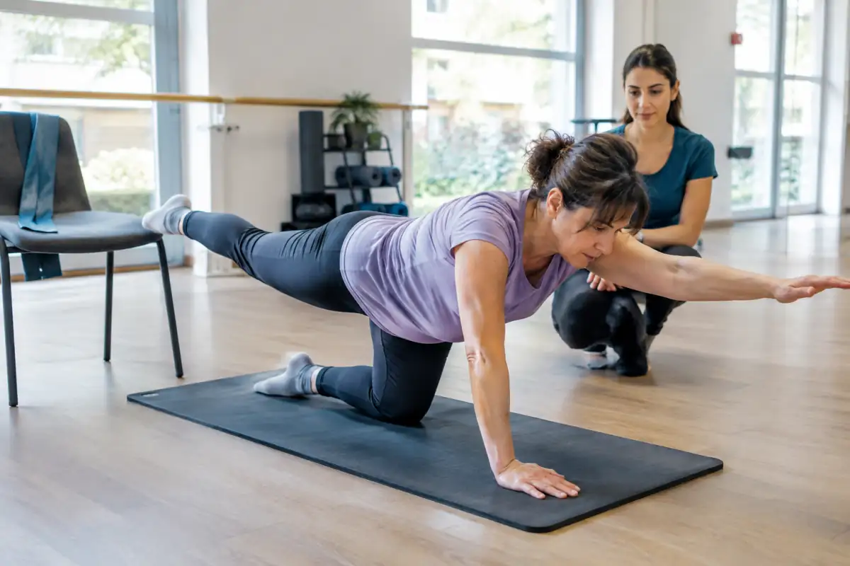 Une pratiquante réalise un exercice de renforcement postural à quatre pattes sur un tapis, sous le regard d’un encadrant.