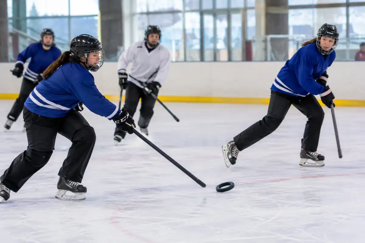 Joueuses de ringette sur la glace, l’une passe un anneau avec une crosse droite sans palette tandis qu’une partenaire se démarque.