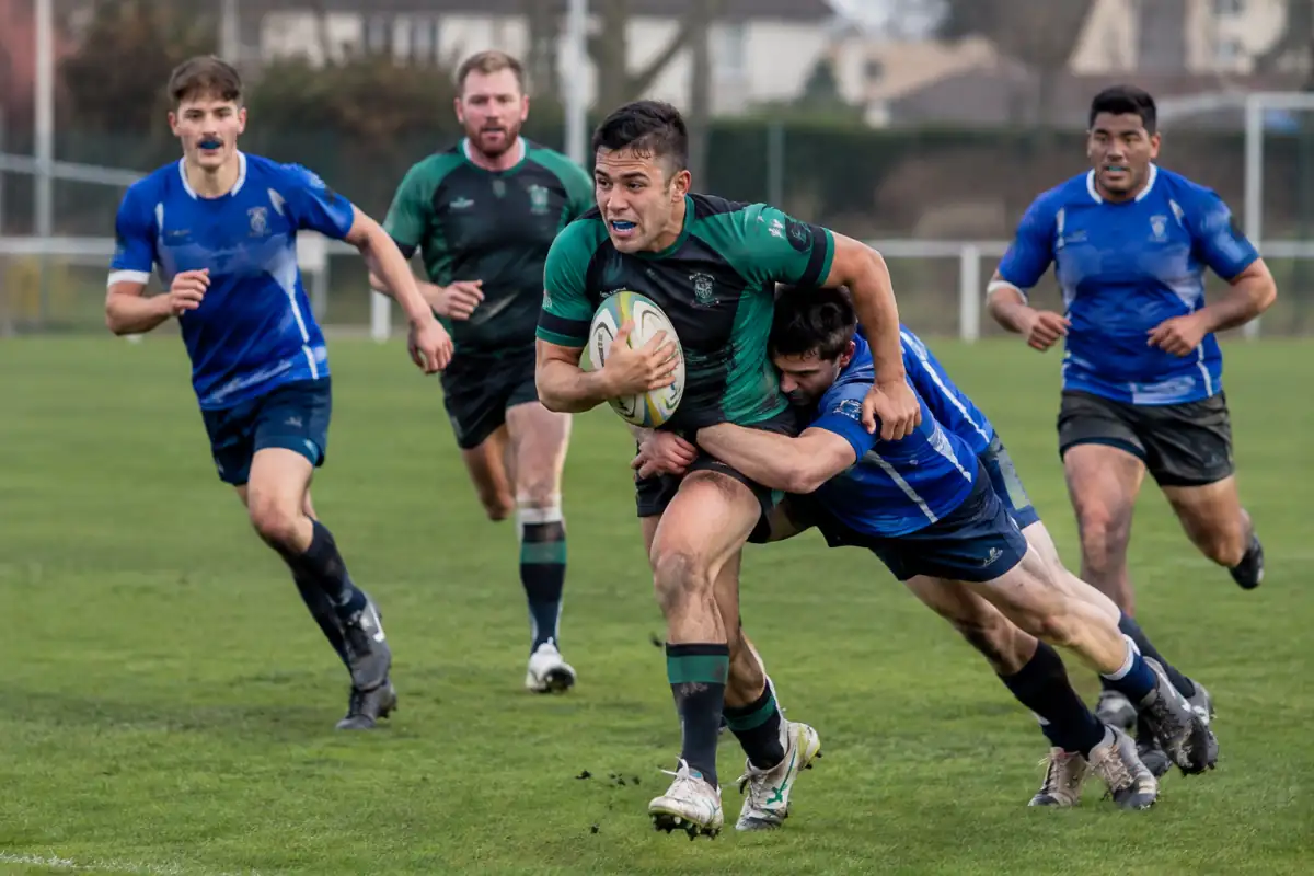 Action de rugby à 9 sur un terrain en herbe, avec un porteur de balle soutenu par ses partenaires et un défenseur au plaquage.