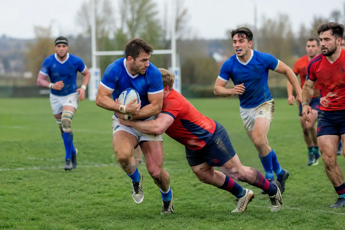Des joueurs de rugby à XV disputent le ballon sur un terrain, avec un porteur soutenu par ses coéquipiers et plaqué par un adversaire.