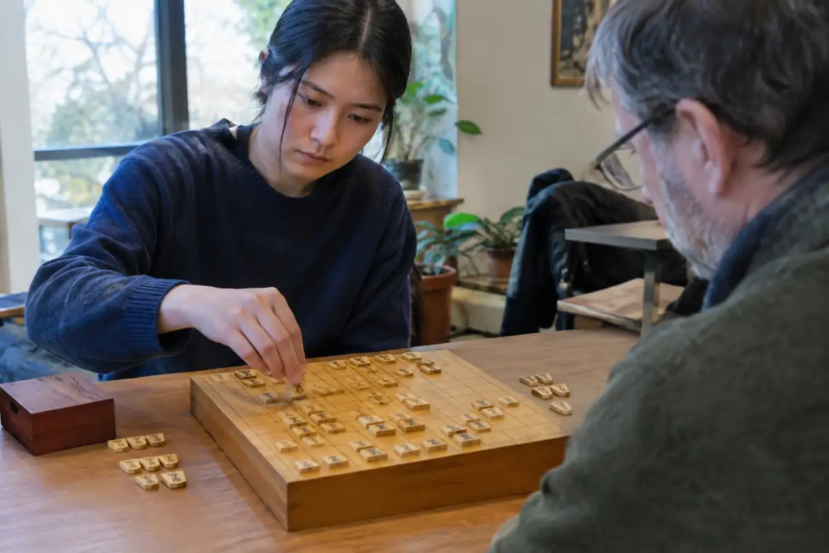 Deux joueurs disputent une partie de shogi sur un plateau en bois, l’un déposant une pièce capturée sur une case libre.