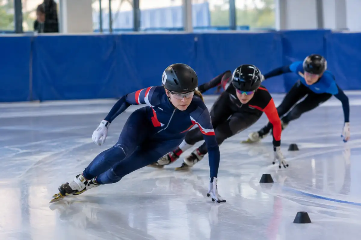 Patineurs de short-track en virage, inclinés sur leurs lames dans une course groupée sur une piste glacée.