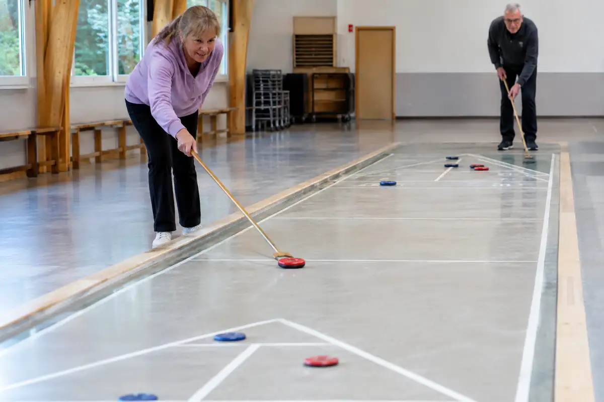 Des joueurs poussent des palets à la queue sur un terrain de shuffleboard au sol marqué de zones de score.