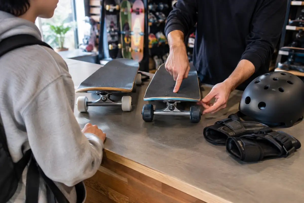 Débutant comparant la largeur de deux skateboards complets, leurs roues et leurs trucks avec un casque et des protections.