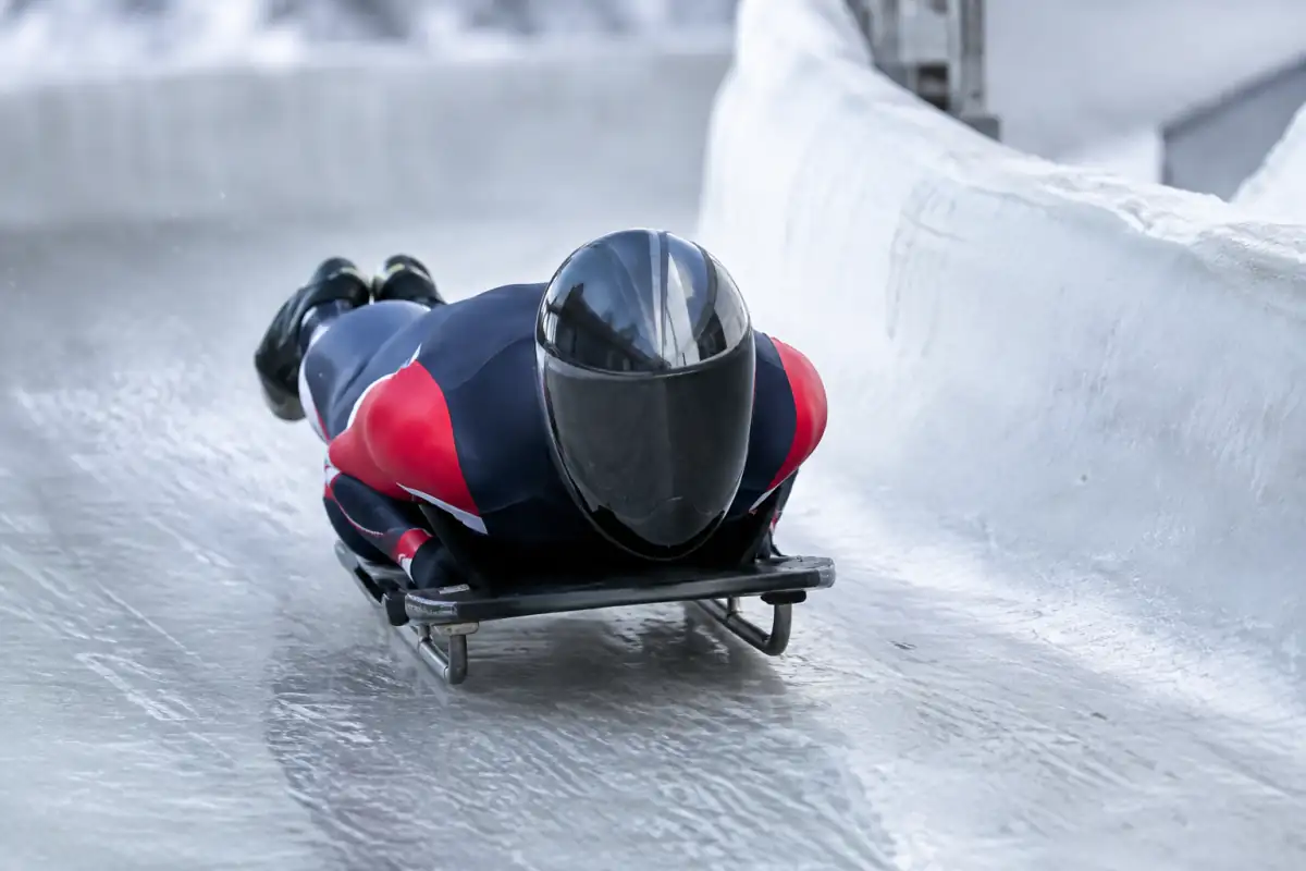 Athlète en casque intégral pilotant un traîneau de skeleton à plat ventre dans un virage glacé.