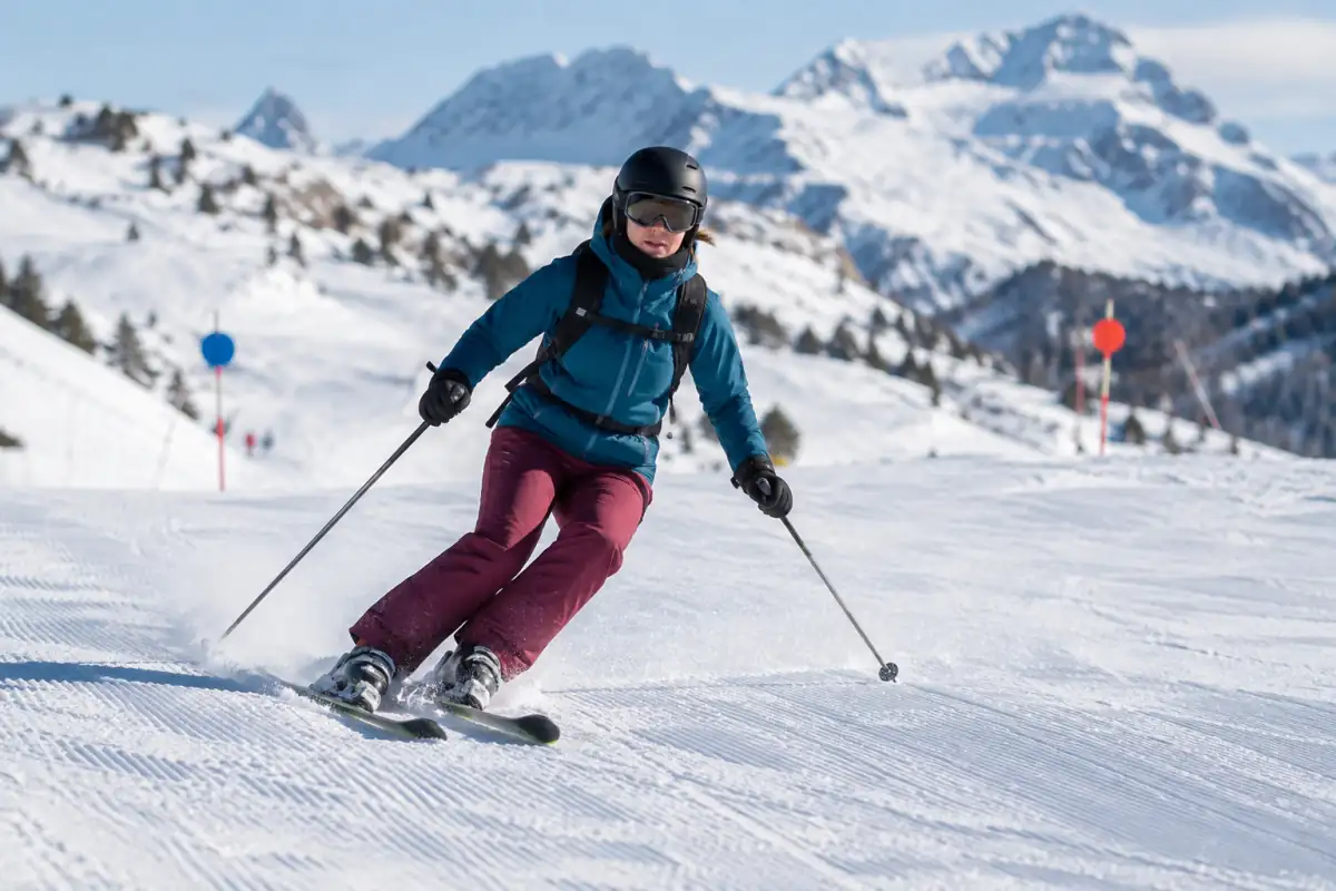 Skieur en virage parallèle sur une piste damée des Alpes françaises, équipé de skis, casque, masque et bâtons.