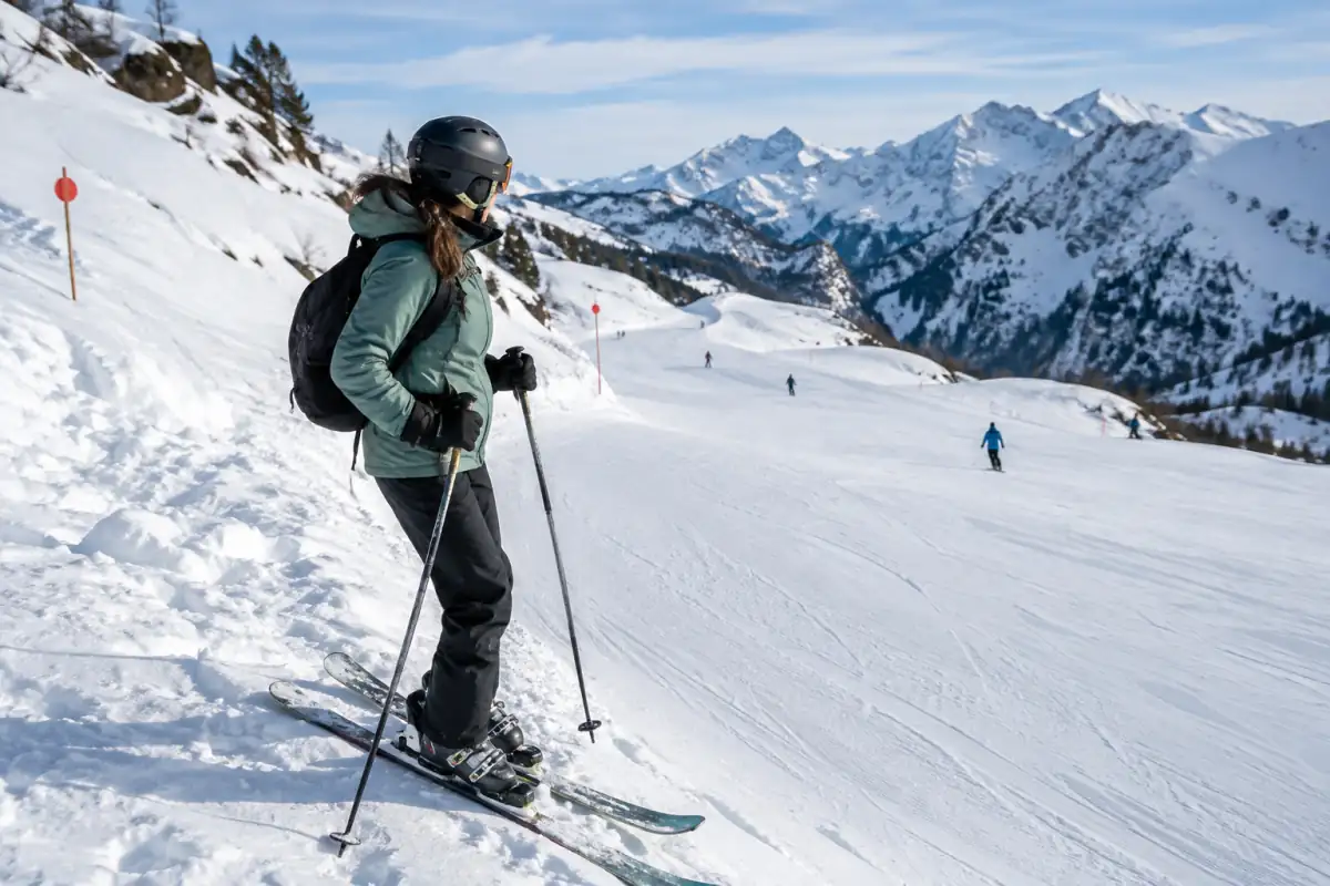 Skieur arrêté en bordure d’une piste, regardant vers l’amont avant de repartir, tandis que d’autres skieurs gardent leurs distances.