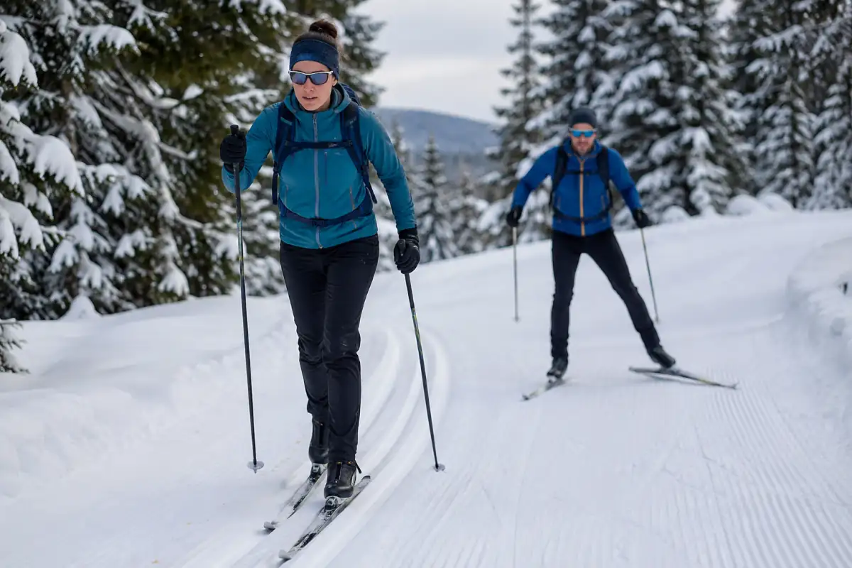 Deux skieurs pratiquent le ski de fond en style classique et skating sur une piste damée dans un paysage enneigé.