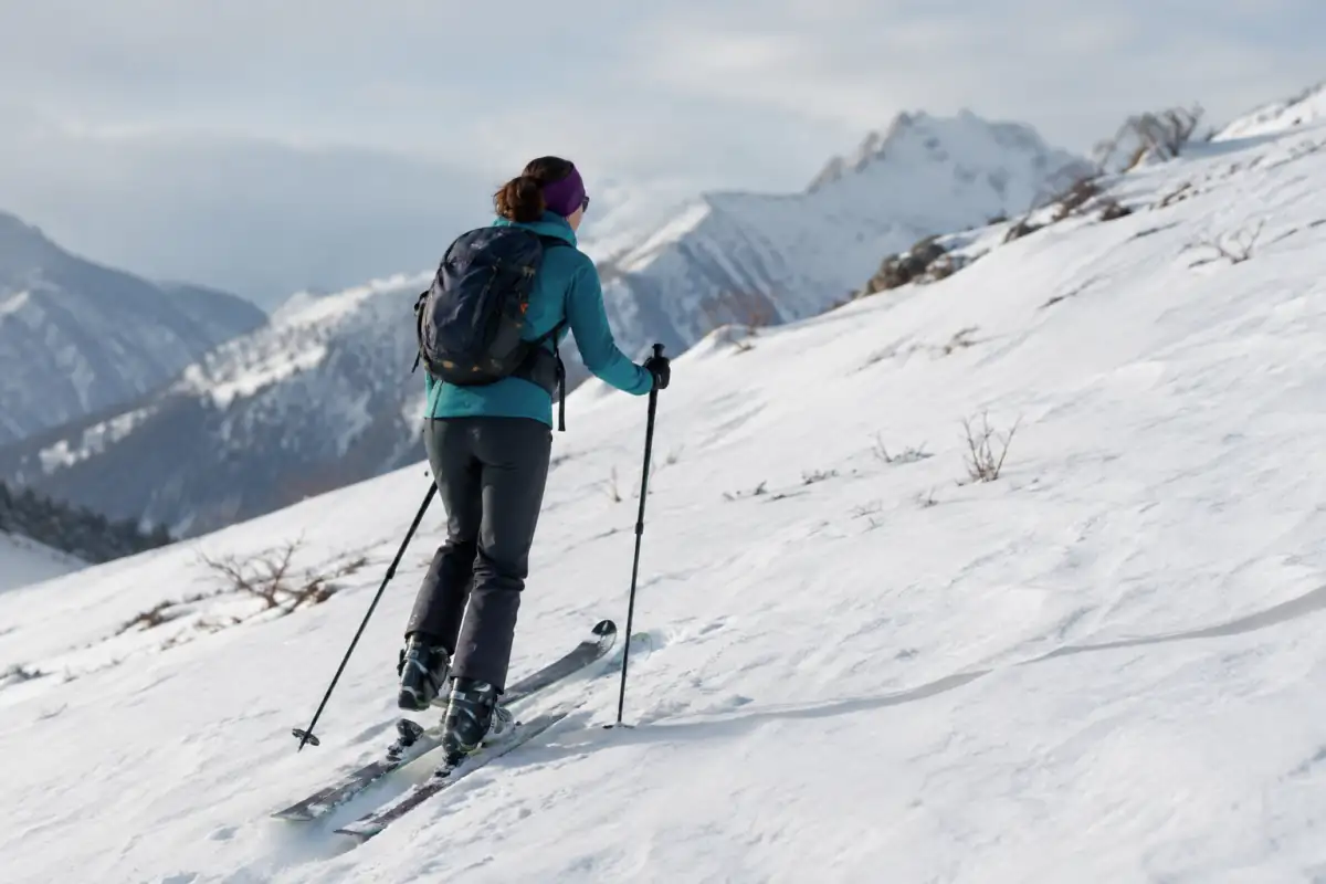 Skieur de randonnée en montée à allure régulière, skis équipés de peaux et bâtons posés dans une trace enneigée.