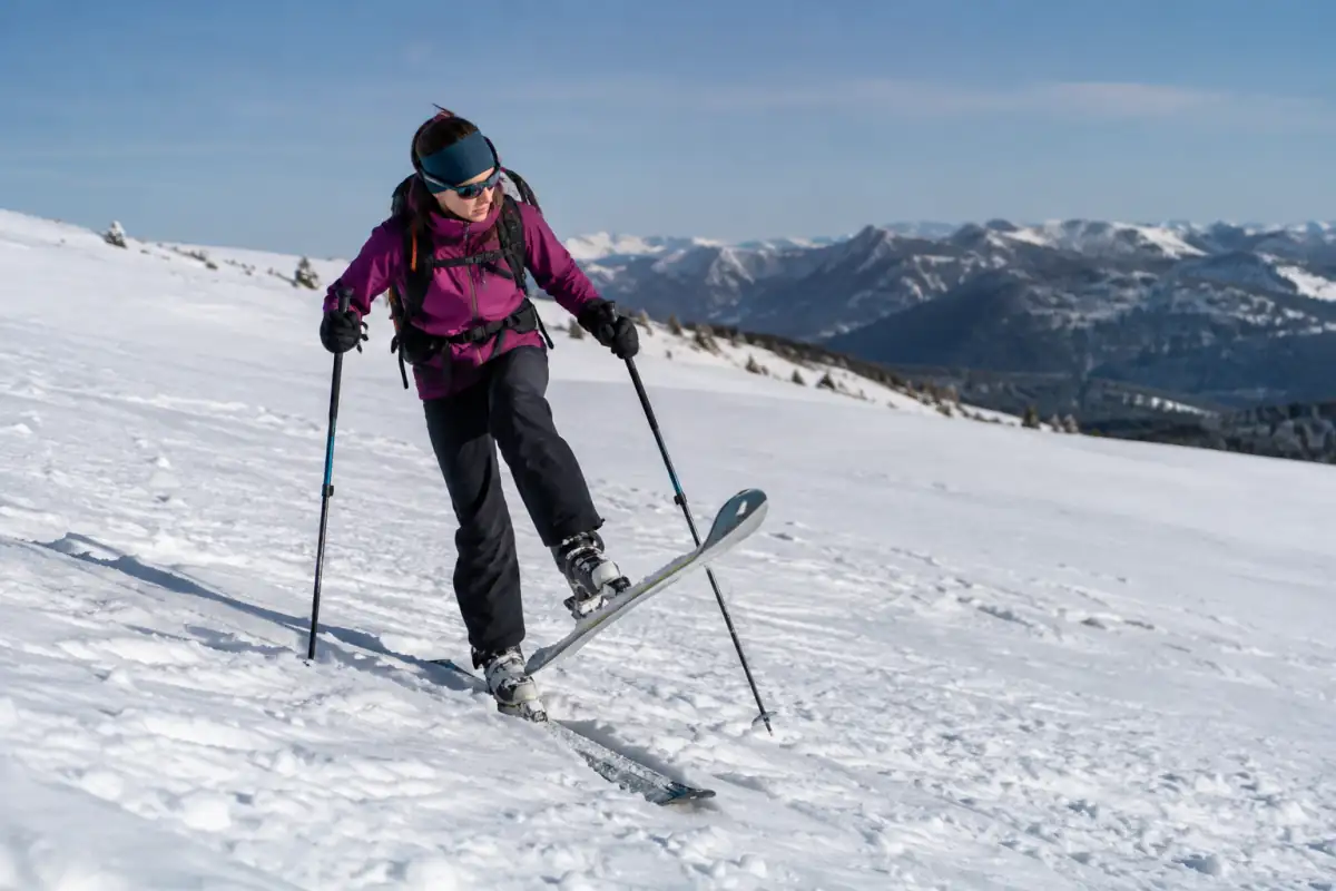 Skieuse en traversée sur une pente douce, pivotant un ski à l’amont avec les peaux en place et les bâtons en appui.