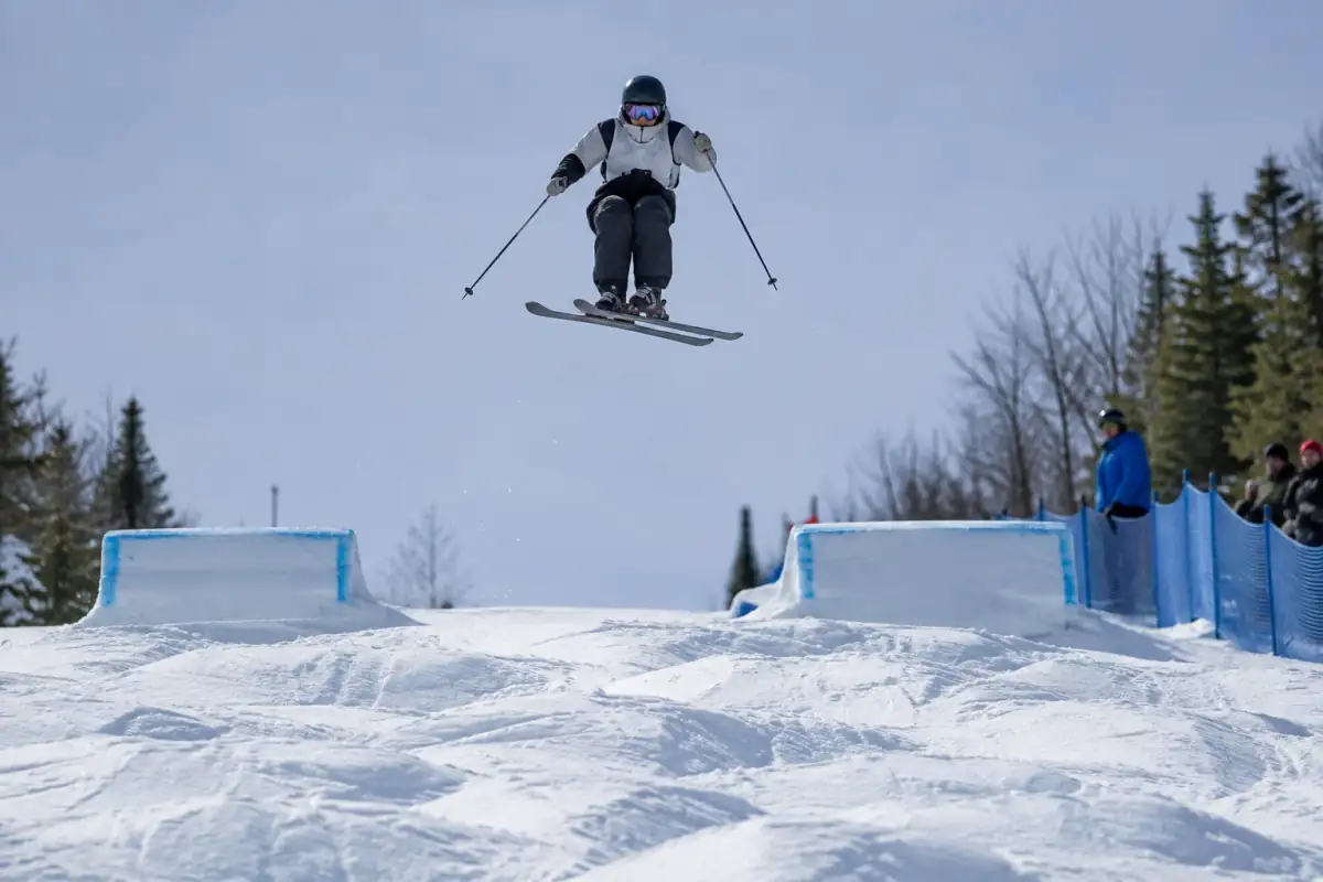 Skieur en compétition au-dessus d’un parcours de bosses, en plein saut, avec une réception maîtrisée et les juges en arrière-plan.