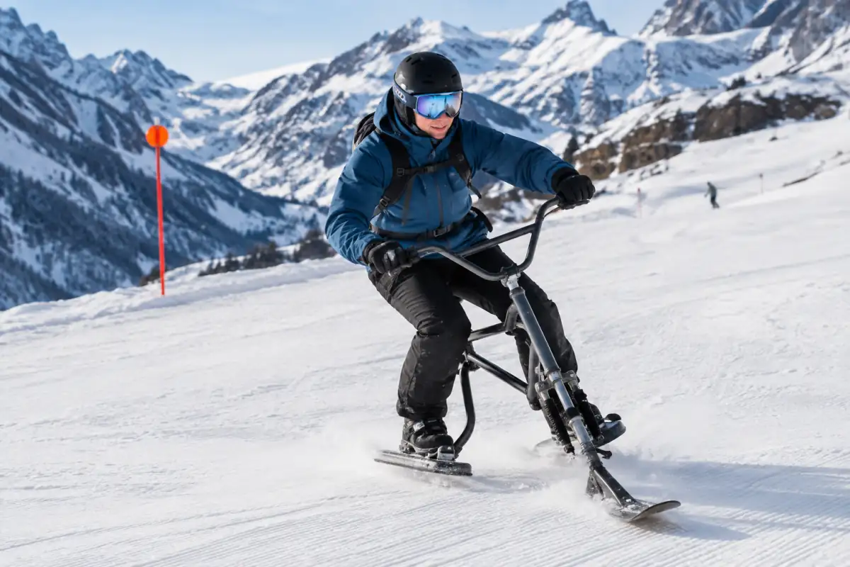 Un pratiquant descend une piste enneigée en skibob, guidon en main, avec casque et petits skis fixés aux pieds.