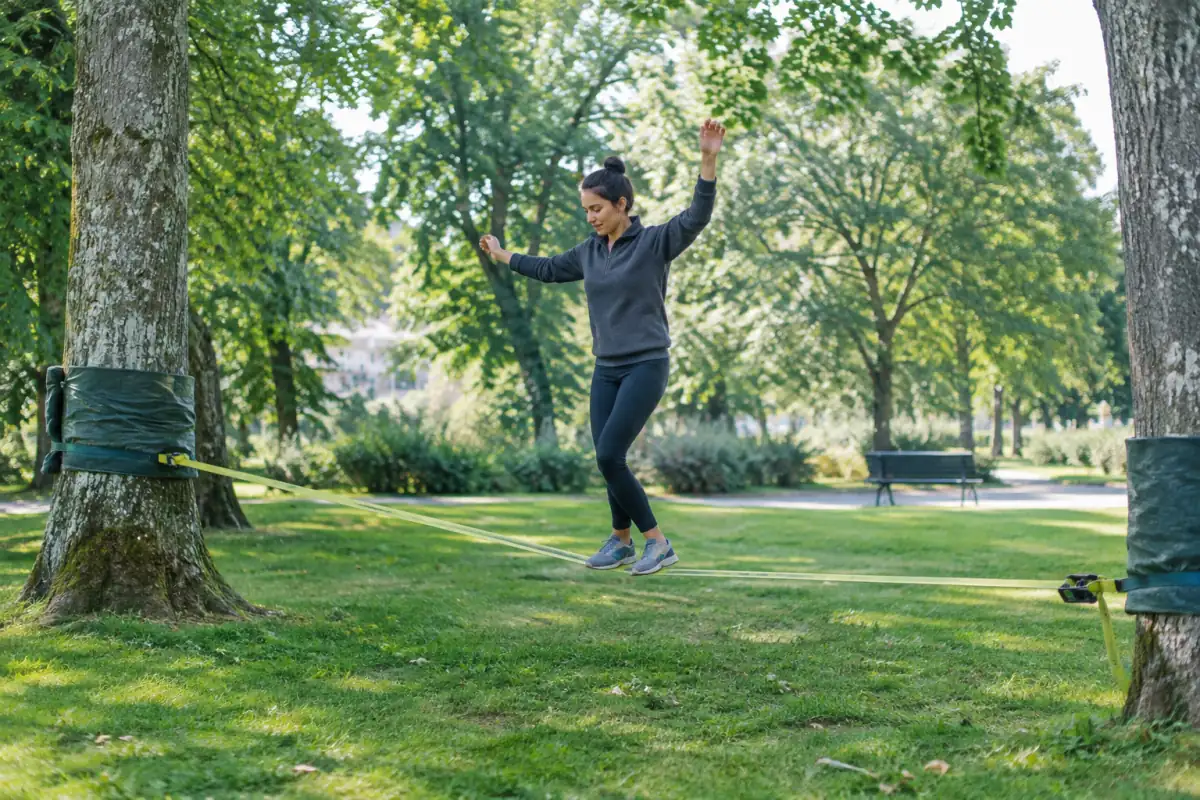 Pratiquant débutant en équilibre sur une slackline basse protégée entre deux arbres dans un parc dégagé.