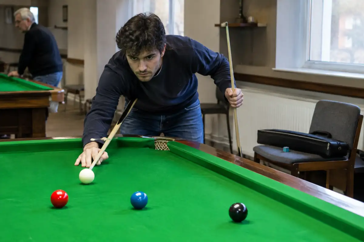 Un débutant compare deux queues de snooker près d’une table, avec une craie et un étui posés à proximité.
