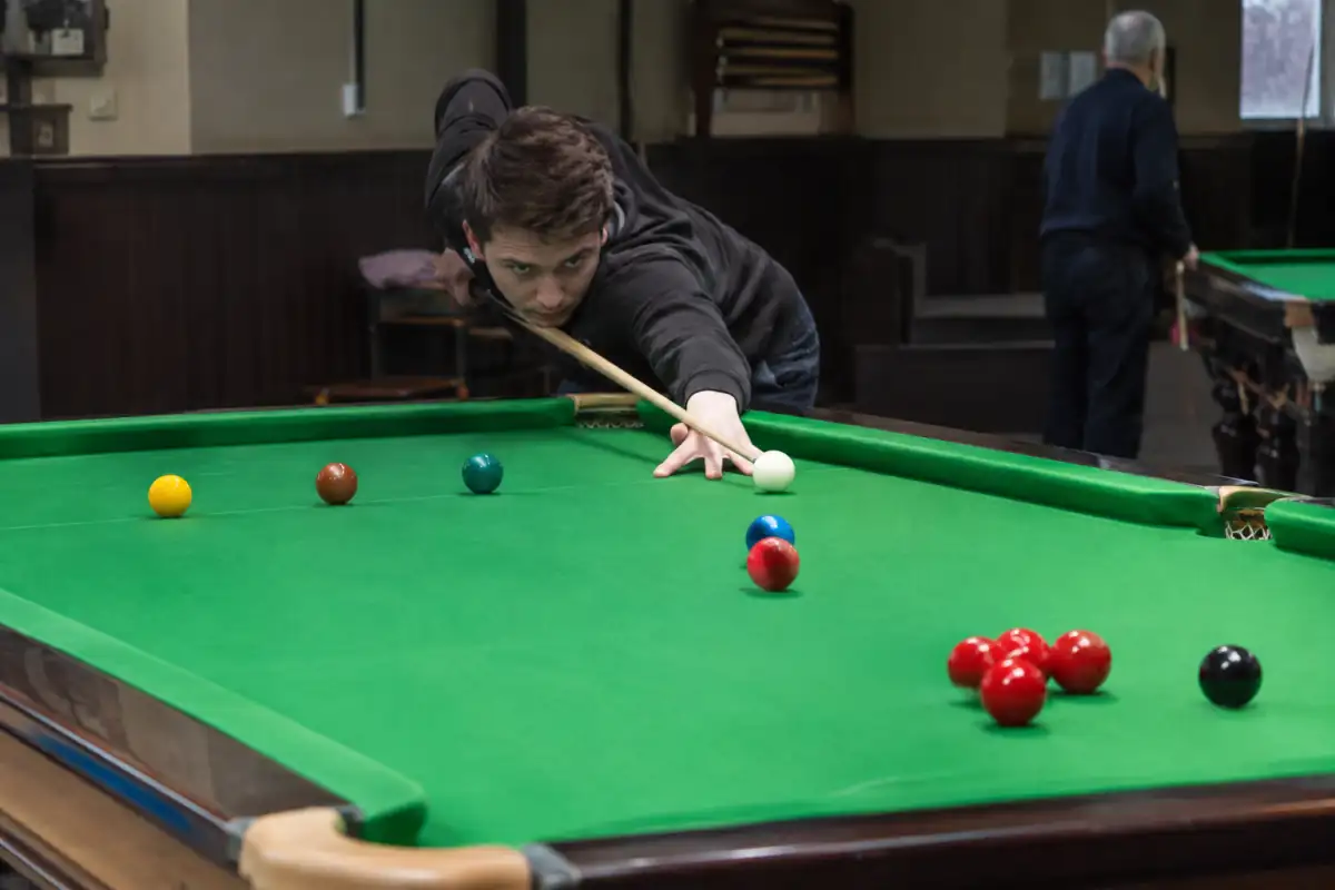 Joueur de snooker préparant une safety près de la bande, avec la blanche masquée par une bille de couleur devant une rouge.
