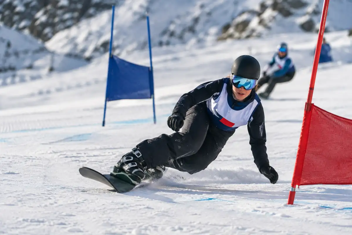 Un snowboardeur en équipement de course carve entre les portes d’un slalom parallèle sur une piste enneigée des Alpes.
